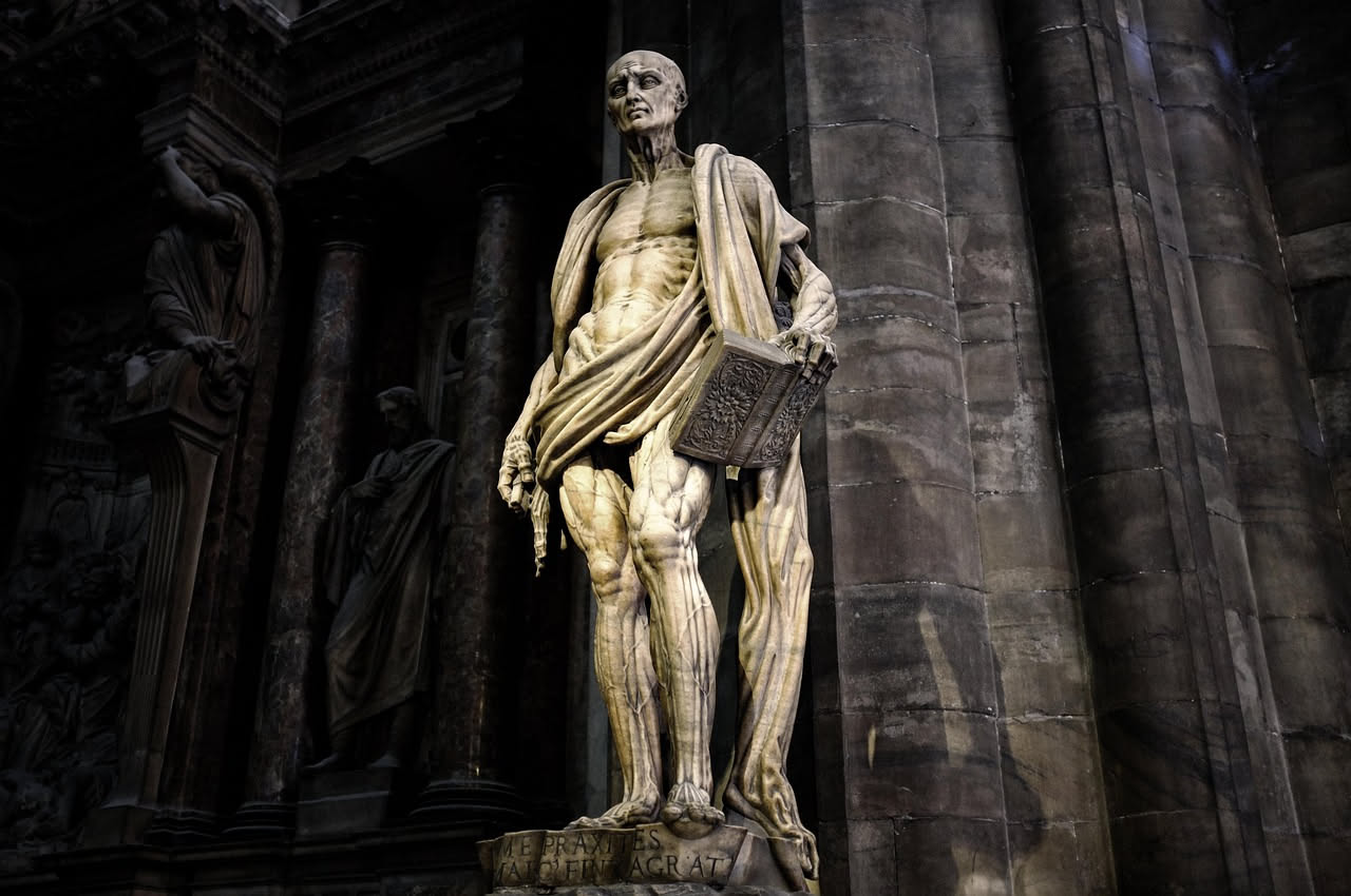 San Girolamo anziano con barba bianca, in veste monastica scura, intento a scrivere con calamo su pergamena in una cella monastica medievale, con libri e manoscritti sugli scaffali sullo sfondo, luce